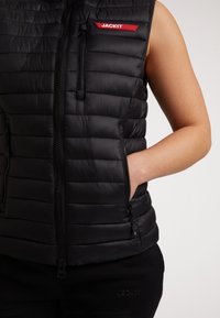 Gilet imbottito nero con zip frontale, tasche laterali e cuciture orizzontali; presenta un'etichetta logo rossa. Tessuto morbido e lucido.
