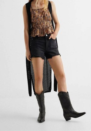 Femme portant des bottes de cowboy noires, un short en denim noir, un haut sans manches marron à motifs, et un long gilet noir en maille ajourée.