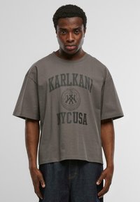 Pelēks kokvilnas T-krekls ar brīvu piegriezumu, īsām piedurknēm, uz kura priekšpuses ir izteiksmīgi melni burti "KARLKANI NYC USA" un apļveida emblēmas dizains.