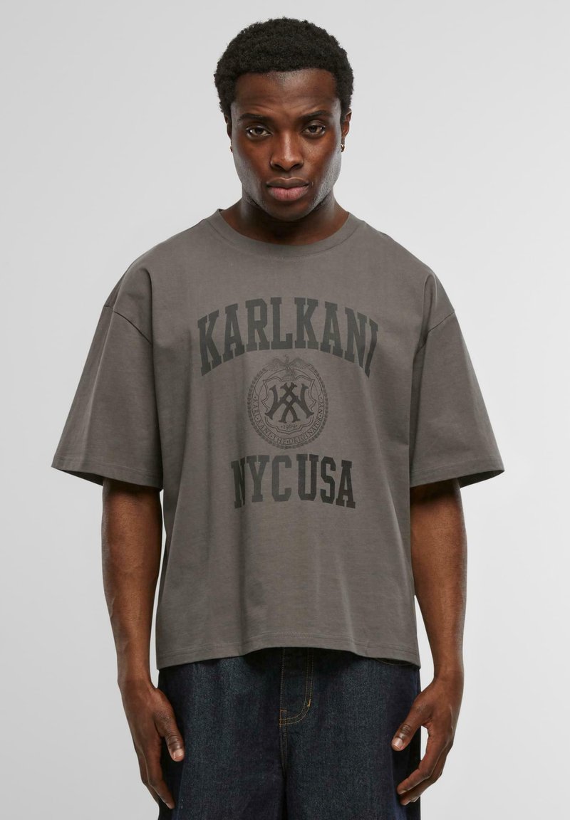Pelēks kokvilnas T-krekls ar brīvu piegriezumu, īsām piedurknēm, uz kura priekšpuses ir izteiksmīgi melni burti "KARLKANI NYC USA" un apļveida emblēmas dizains.