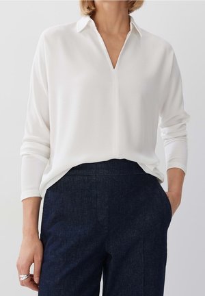 Kobieta w białej bluzce z długim rękawem i dekoltem w serek, schowanej w ciemnoniebieskie spodnie z wysokim stanem, z jedną ręką w kieszeni.