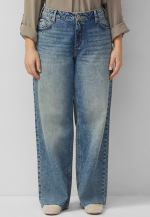 Person in hellblauen Jeans mit weitem Bein und beigen, strukturierten Slippern, die vor einem schlichten grauen Hintergrund steht.