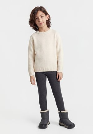 Bambino con capelli castano chiaro fino alle spalle, indossa un maglione lavorato a maglia color crema, leggings grigio scuro e stivali invernali grigi, in piedi su uno sfondo bianco.