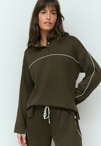 Pull-over marron avec un col montant, boutons-pression et coutures blanches. Accompagné de pantalons assortis avec une taille à cordon et des accents noirs.