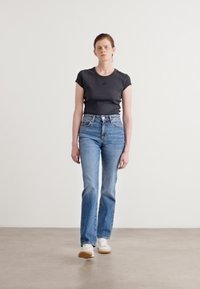 Svart t-skjorte med korte ermer, høyimponerte lyseblå jeans med rett ben, og hvite joggesko, satt opp mot en enkel bakgrunn.