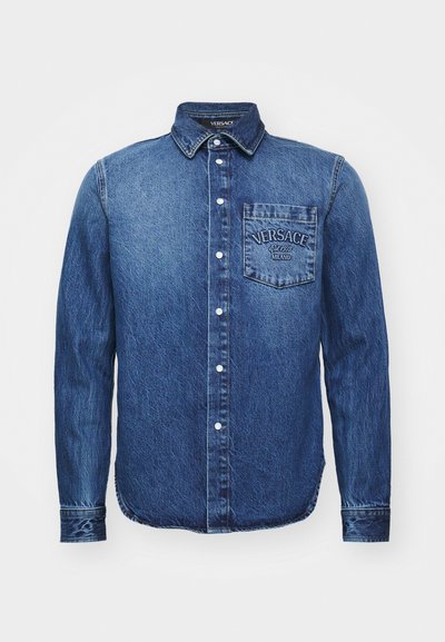 Camicia in denim blu scuro con maniche lunghe, colletto button-down, taschino anteriore con logo "Versace" e dettagli di cucitura in evidenza.