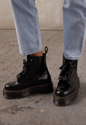Person trägt schwarze Leder-Schnürstiefel mit dicken Sohlen und gelben Nähten, kombiniert mit hellblauen Jeans auf Betonboden.
