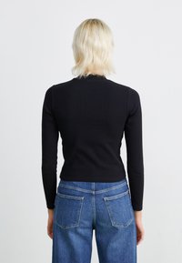 Svart langermet topp med tettsittende design, rillet tekstur og høy hals, kombinert med høyhalsede blå denimjeans med baklommer.