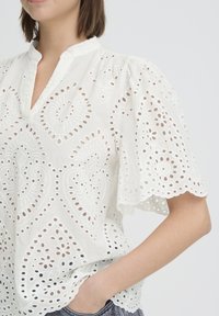 Femme portant un chemisier blanc en broderie anglaise avec manches bouffantes et bords festonnés, main dans la poche, sur fond uni.