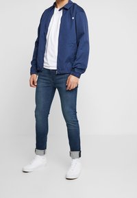 Mörkblå lätt jacka med krage, kombinerad med en vit t-shirt, mörkblå jeans och vita sneakers. Jackan har en logotyp på bröstet.