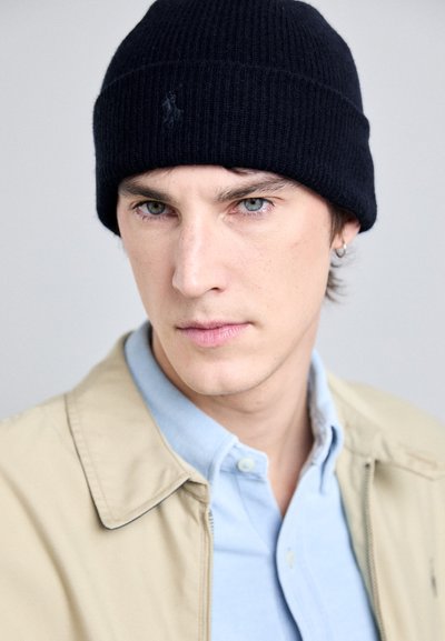 Gorro de punto negro con acanalado, camisa de color azul claro con cuello y chaqueta beige. Enfocarse en la cara y rasgos distintivos como la oreja perforada y la expresión.