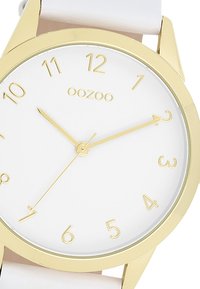 Montre dorée avec un cadran blanc, grands index horaires dorés et aiguilles dorées fines. Attachée à un bracelet blanc pour un look épuré et moderne.