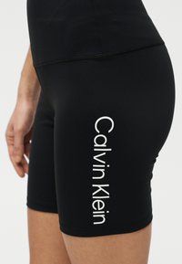 Κοντινή λήψη μαύρων κοντών εφαρμοστών σορτς με κάθετο λευκό λογότυπο "Calvin Klein" στον μηρό που φοριέται από άτομο.