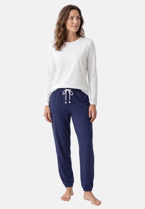 Kobieta ubrana w białą, długorękawową bluzkę i granatowe spodnie z troczkami, stojąca boso na jednolitym tle.