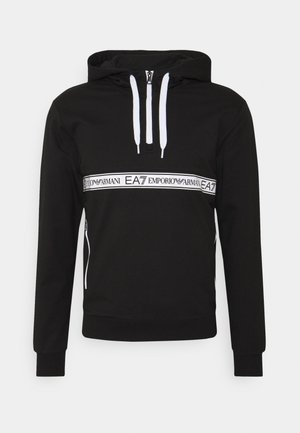 Hoodie noir en tissu doux, avec une bande de logo blanche sur la poitrine, fermeture éclair à l'avant et capuche avec cordon de serrage. Poches latérales visibles.