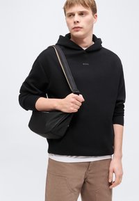 Νεαρός άνδρας που φοράει μαύρο φούτερ με κουκούλα BOSS και μπεζ παντελόνι, κρατώντας ιμάντα μαύρης τσάντας χιαστί στον ώμο του, μπροστά σε απλό φόντο.