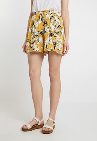 Žluté květinové šortky se sbíraným pasem, se vzorem černých a bílých květin na strukturované látce. Nosené s bílými sandály.