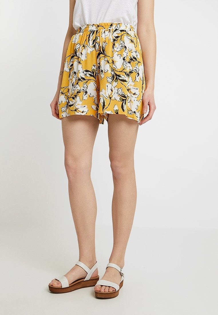 Žluté květinové šortky se sbíraným pasem, se vzorem černých a bílých květin na strukturované látce. Nosené s bílými sandály.