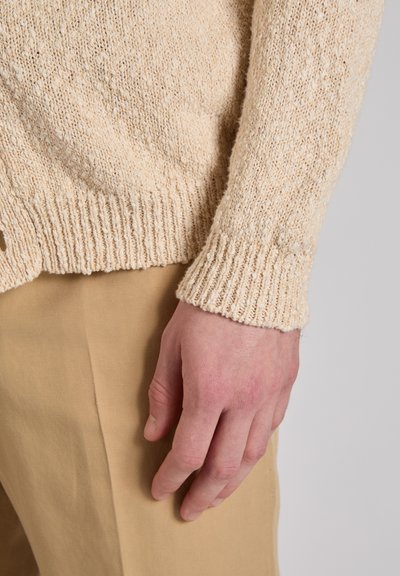 Knit sweater in off-white with a textured pattern and ribbed cuffs. Hand positioned near light beige fabric of pants.