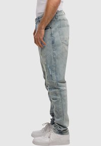 Jeans de mezclilla de lavado claro con detalles desgastados, corte ajustado, bolsillos laterales y un ligero desgaste. Llevados con zapatillas blancas.