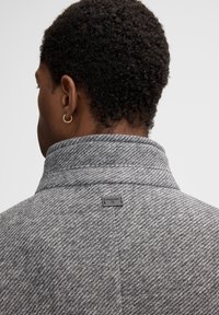 Cappotto grigio a motivo a spina di pesce con colletto alto e etichetta sul retro. Il tessuto sembra avere una texture e è lavorato a maglia, offrendo calore.