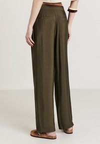 Frau trägt lockere, olivgrüne Satin-Hosen mit weitem Bein, hellbraune Mokassins und weiße sowie braune Armreifen, steht mit dem Rücken zur Kamera.