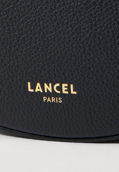 Логото на Lancel Paris върху текстурирана тъмносиня кожа, с позлатени ембосирани букви. Повърхността показва деликатен, естествен модел на зърно.
