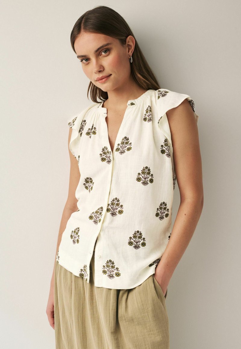 Witte blouse met korte mouwen, versierd met groene en bruine bloemenborduursels, met knopen aan de voorkant en ruffle-mouwen, gecombineerd met beige, flowy broek.