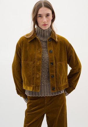 Giacca in velluto a coste marrone con colletto, chiusura a bottone e tasche frontali, indossata sopra un maglione beige strutturato. Pantaloni in velluto a coste marroni abbinati.