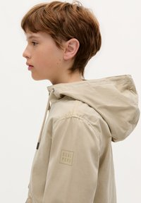 Giacca leggera beige con cappuccio, dotata di cordoncino e impreziosita da un sottile logo "SCALPERS" sulla manica.