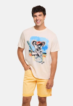 Hombre sonriendo con una camiseta beige con un koala de dibujos animados jugando al béisbol y pantalones cortos amarillos, de pie frente a un fondo liso.