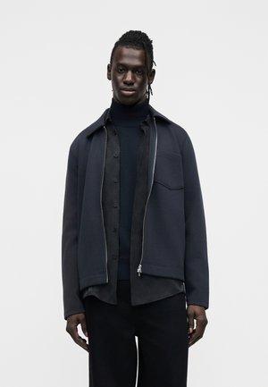 Homme aux cheveux tressés portant une veste superposée noire et bleu foncé, une chemise boutonnée et un pull à col roulé sur un fond gris uni.