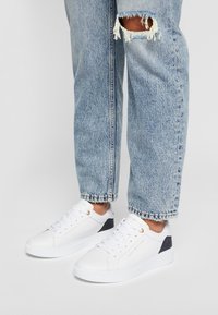 Zapatillas blancas con una superficie lisa, costuras detalladas y ojetes dorados. Acento texturizado en azul marino en el talón. Combinadas con unos vaqueros ligeros desgastados.