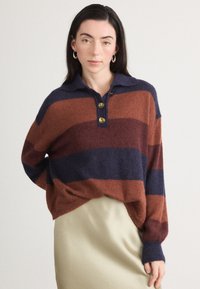 Pull oversized tricoté en ambre, bleu marine et bordeaux rayé. Présente un col, trois boutons dorés et des poignets côtelés.