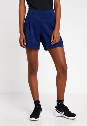 Person iført mørkeblå plisserede shorts, sorte Nike-sneakers med hvide såler og sorte sokker, stående på en hvid gulv.