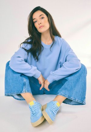Felpa oversize azzurro chiaro, jeans denim a gamba larga e sneakers blu con dettagli gialli. Texture liscia in uno stile casual e rilassato.
