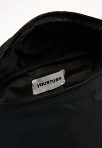 Musta kangaskassi, jonka sisällä on sileä pinta, ja siinä on pieni valkoinen etiketti, jossa lukee "YOURTURN". Vetoketju yläreunassa.