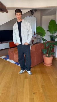 Šedá košile s knoflíky přes černé tričko, černé kalhoty a stříbrné tenisky, v místnosti s rostlinami a hnědou pohovkou.