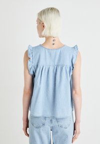 Svetlo modra denim brezrokavnik z nabranimi rameni, nabiranim hrbtom in sproščenim krojem. Dopolnjen z usklajenimi denim hlačami.