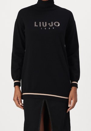 Pull noir à col roulé manches longues avec bordure beige et texte "LIU·JO 1995" en strass sur la poitrine, porté avec une jupe noire fendue devant.