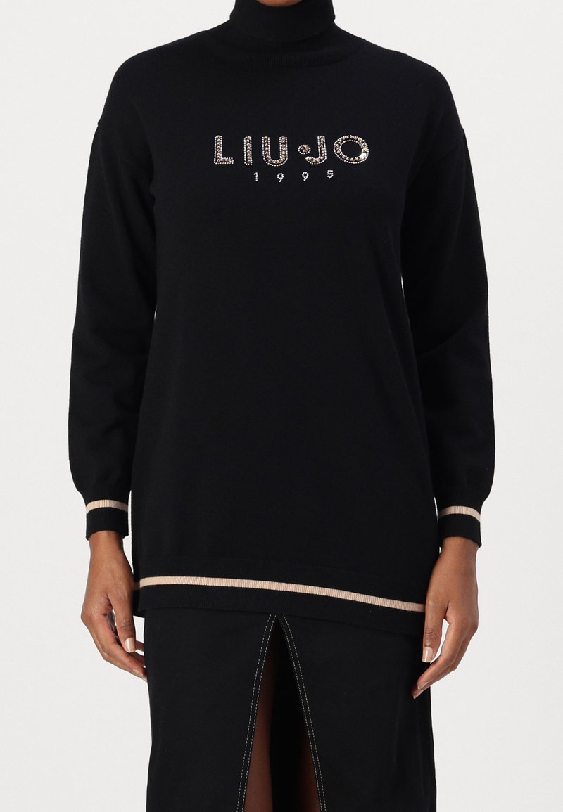 Pull noir à col roulé manches longues avec bordure beige et texte "LIU·JO 1995" en strass sur la poitrine, porté avec une jupe noire fendue devant.