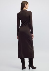 Langärmliges, tailliertes braunes Kleid aus glattem Stoff mit einer eleganten Silhouette und seitlichem Raffung, kombiniert mit knielangen Stiefeln aus Wildleder.