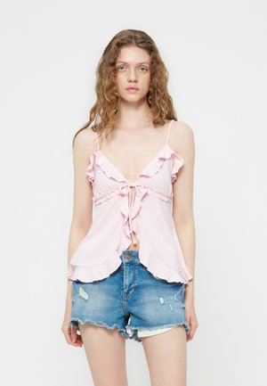 Jeune femme aux cheveux bouclés portant un haut sans manches rose clair à volants et un short en jean bleu déchiré, debout devant un fond blanc.