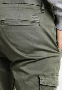Olivgrüne Cargo-Hosen aus glattem Stoff, mit einer Gesäßtasche und einem Guess-Logo-Patch in der Nähe des Bundes.
