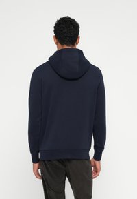 Ναυτικό μπλε φούτερ με ρυθμιζόμενο κορδόνι, μαλακή υφή, χαλαρή εφαρμογή, ριμπέ ύφανση στις μανσέτες και το στρίφωμα. Φορεμένο πάνω από ριγέ παντελόνια από κορντούρα.