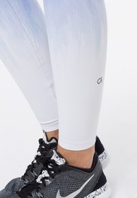 Nohy oblečené v legínách s bílým až světle modrým gradientem a černých běžeckých botách Nike s bílými solemi s rozstřikovaným vzorem na bílém pozadí.