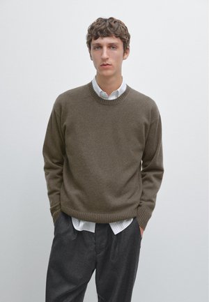 Donkergroene trui met een ronde hals en geribbelde zoom, gedragen over een wit gestreept shirt. In combinatie met losse, donkergrijze pantalons.