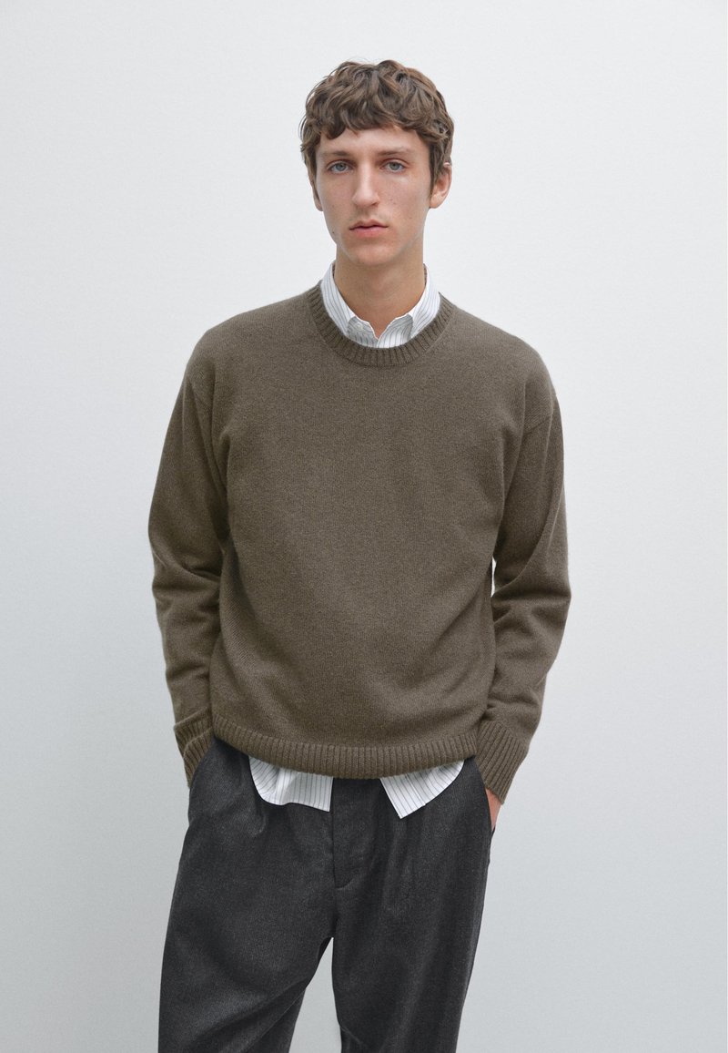 Donkergroene trui met een ronde hals en geribbelde zoom, gedragen over een wit gestreept shirt. In combinatie met losse, donkergrijze pantalons.