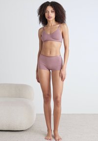 Mauve bra and matching briefs set, featuring a smooth texture, soft fabric, and minimalist design. Model stands on a light carpet.