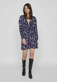 Vestido floral envolvente en negro con patrones abstractos en azul, rosa y blanco. Mangas largas, escote en V y cintura ceñida, combinado con botas negras brillantes.